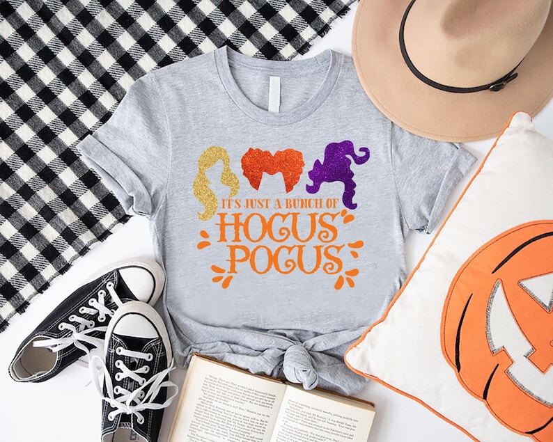 Op de afbeelding: Een grijs T-shirt met een afbeelding van drie heksensilhouetten en de tekst "It's just a bunch of Hocus Pocus".