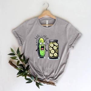 May include: A gray t-shirt featuring a cartoon pickle with a shocked expression next to a jar of pickle slices. The pickle has a green body, black eyes, and a wide open mouth. The jar is filled with yellow-green pickle slices.