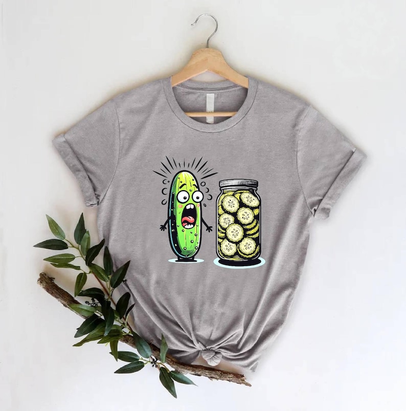 May include: A gray t-shirt featuring a cartoon pickle with a shocked expression next to a jar of pickle slices. The pickle has a green body, black eyes, and a wide open mouth. The jar is filled with yellow-green pickle slices.