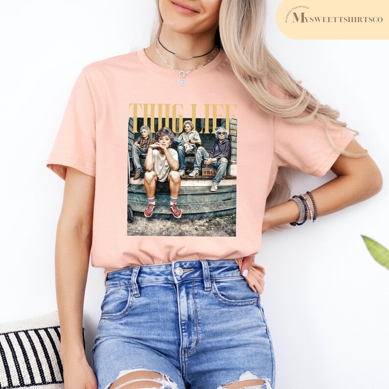 May include: A pink t-shirt with a graphic print of four people sitting on a stoop. The graphic includes the text "THUG LIFE".