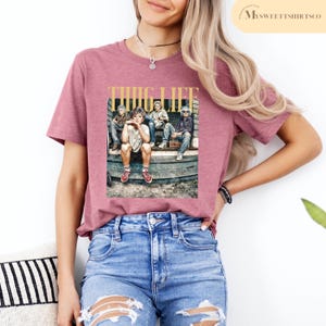 May include: A pink t-shirt with a graphic print of four people sitting on a porch. The graphic has the text "THUG LIFE" in gold letters.