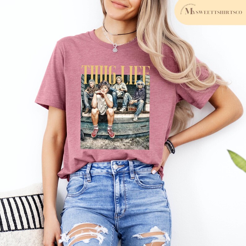 May include: A pink t-shirt with a graphic print of four people sitting on a porch. The graphic has the text "THUG LIFE" in gold letters.