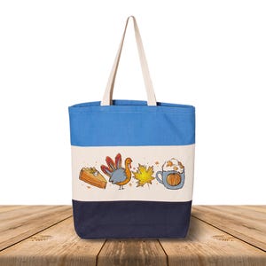 May include: A canvas tote bag with a blue top, cream middle, and navy bottom. The bag features a Thanksgiving-themed illustration with a slice of pie, a turkey, a maple leaf, and a pumpkin spice latte.