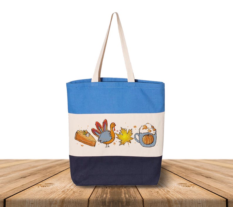 May include: A canvas tote bag with a blue top, cream middle, and navy bottom. The bag features a Thanksgiving-themed illustration with a slice of pie, a turkey, a maple leaf, and a pumpkin spice latte.