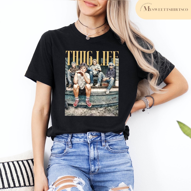 May include: A black t-shirt with the text 'THUG LIFE' in yellow letters. The t-shirt features a photo of four people sitting on a wooden bench. The people are wearing casual clothing and are looking at the camera. The t-shirt is being worn by a person with blue jeans.