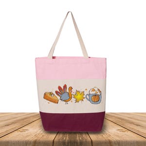 May include: A pink, cream, and burgundy tote bag with cream-colored handles. The bag features a colorful illustration of a slice of pie, a turkey, a maple leaf, and a pumpkin spice latte. The bag is made of canvas.