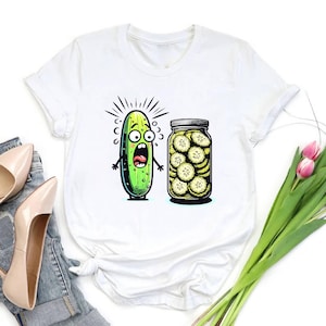 May include: White t-shirt featuring a cartoon pickle with a shocked expression next to a jar of pickle slices. The pickle is green with black outlines and a wide-open mouth. The jar is filled with yellow-tinged pickle slices.