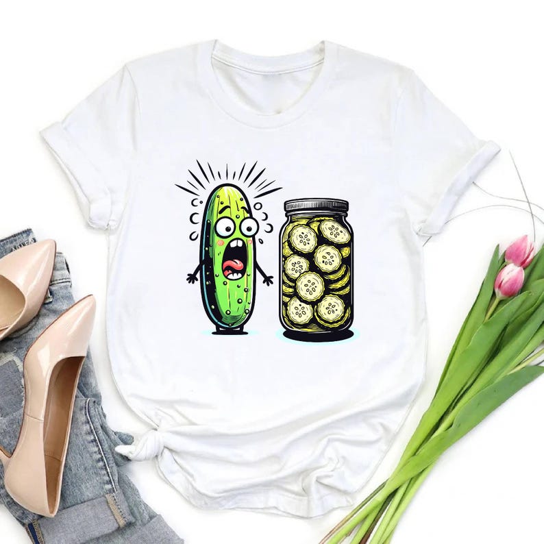 May include: White t-shirt featuring a cartoon pickle with a shocked expression next to a jar of pickle slices. The pickle is green with black outlines and a wide-open mouth. The jar is filled with yellow-tinged pickle slices.
