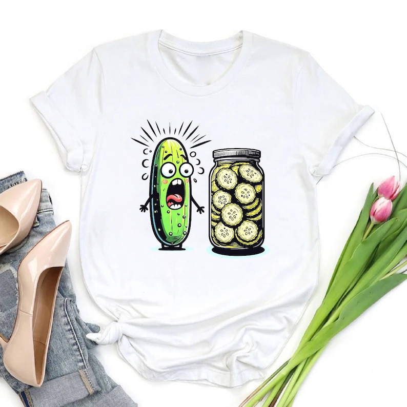 May include: White t-shirt featuring a cartoon pickle with a shocked expression next to a jar of pickle slices. The pickle is green with black outlines and a wide-open mouth. The jar is filled with yellow-tinged pickle slices.