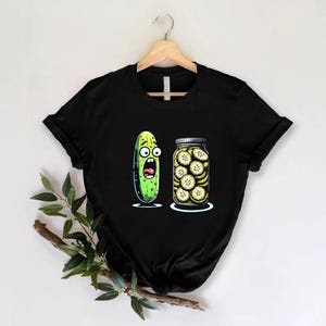 May include: Black t-shirt featuring a cartoon pickle with a shocked expression next to a jar of sliced pickles. The pickle is green with large eyes and an open mouth. The jar is clear with a silver lid.