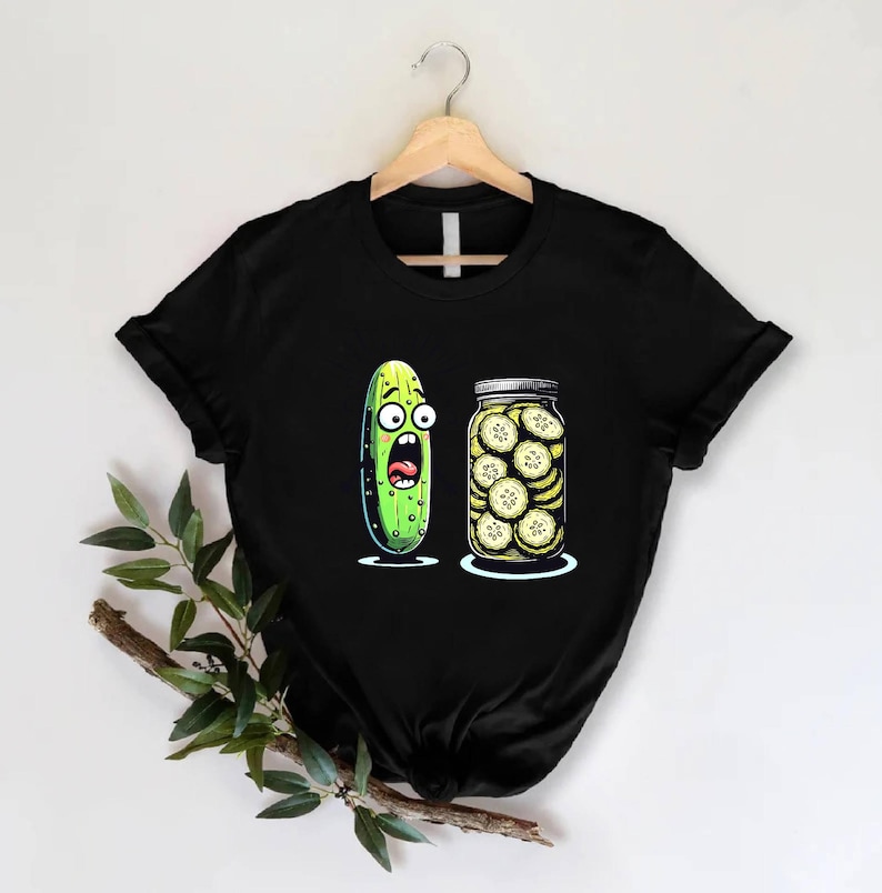 May include: Black t-shirt featuring a cartoon pickle with a shocked expression next to a jar of sliced pickles. The pickle is green with large eyes and an open mouth. The jar is clear with a silver lid.