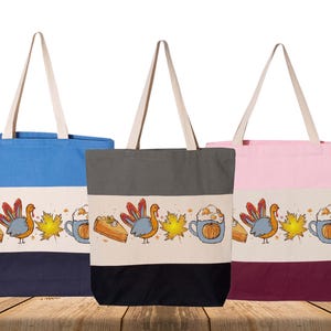 May include: Three canvas tote bags in blue, gray, and pink, each with a cream-colored top and bottom. A Thanksgiving-themed design is printed on a cream band, featuring a turkey, pumpkin spice latte, and pie.