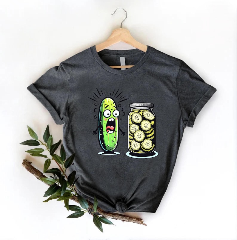 May include: Dark gray t-shirt featuring a cartoon pickle with a shocked expression next to a jar of sliced pickles. The pickle is green with black eyes and a wide open mouth. The jar is clear with a silver lid.