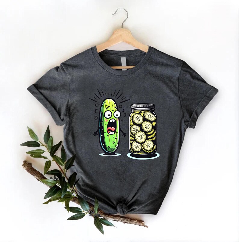 May include: Dark gray t-shirt featuring a cartoon pickle with a shocked expression next to a jar of sliced pickles. The pickle is green with black eyes and a wide open mouth. The jar is clear with a silver lid.