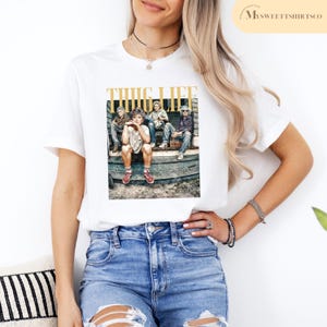 May include: White t-shirt with a graphic print of four people sitting on a porch. The graphic is in black and white and the text "THUG LIFE" is in gold letters.