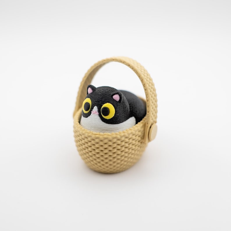 May include: A small, three-dimensional figurine of a black and white cat with large yellow eyes and pink ears, sitting inside a tan, textured basket with a rounded handle. The cat has a white chest and face, and the basket features a woven pattern.