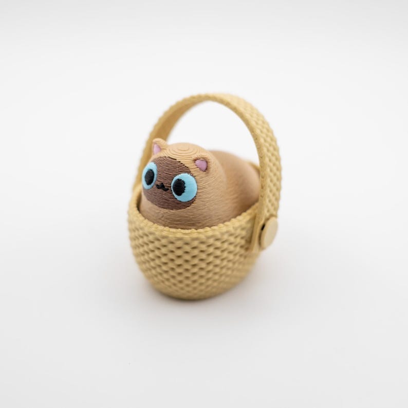 May include: A small, tan, 3D-printed figurine of a cat with a dark brown face and bright blue eyes, sitting inside a light beige, textured, woven-style basket with a single arched handle attached by small circular side fasteners.