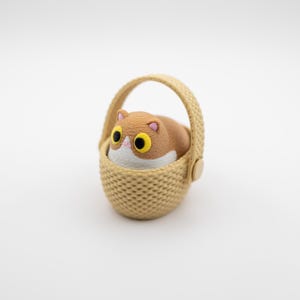 May include: A small figurine of a brown and white cat with large yellow eyes sitting inside a beige woven-style basket. The cat has a rounded body, pink inner ears, and a white patch on its face. The basket features a textured pattern.