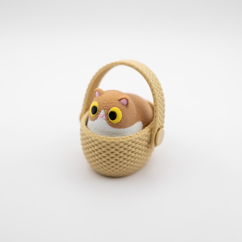 May include: A small figurine of a brown and white cat with large yellow eyes sitting inside a beige woven-style basket. The cat has a rounded body, pink inner ears, and a white patch on its face. The basket features a textured pattern.