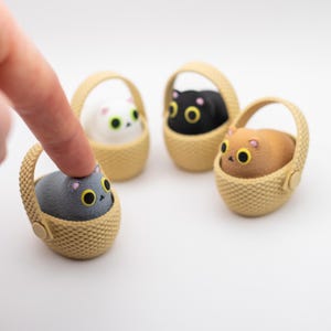 May include: Four small, rounded cat figurines in individual beige woven baskets. The cats are colored gray, white, black, and tan, each featuring large yellow eyes with black pupils and small pink ears. The figurines are arranged on a white surface.
