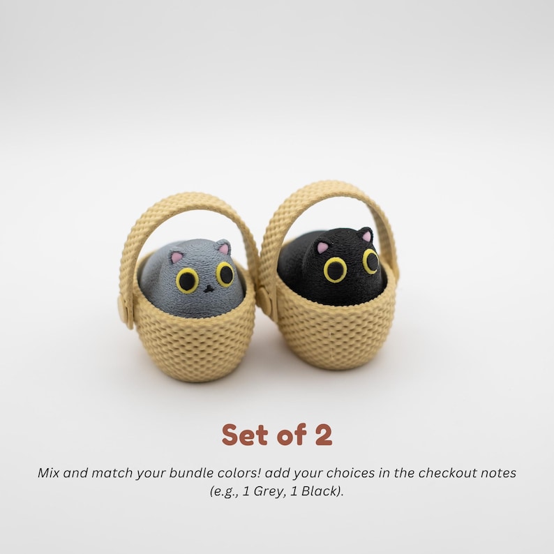 May include: A set of two small figurines, one grey and one black, each shaped like a cat with large yellow eyes and pink ears, sitting inside a beige woven basket. The text below reads: Set of 2. Mix and match your bundle colors! add your choices in the checkout notes (e.g., 1 Grey, 1 Black).