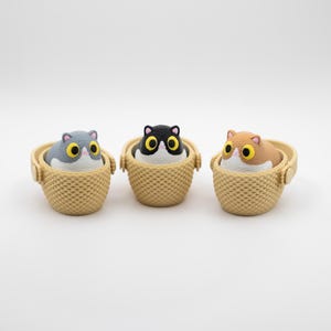 May include: Three small figurines of cats with large yellow eyes, each sitting inside a textured beige basket with a handle. The cats are colored grey, black and white, and light brown, all featuring white chest patches and pink noses.