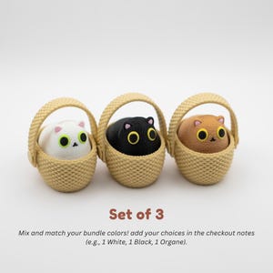 May include: A set of three small cat figurines, each sitting inside a tan woven-texture basket. The cats are white, black, and brown, all featuring large yellow eyes and small pink ears. Text below reads: Set of 3. Mix and match your bundle colors!