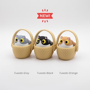 May include: Three small figurines of cats with large yellow eyes, each sitting in a beige woven basket. The cats are colored grey, black, and orange. Above the baskets is a red banner with the text NEW!. Below are the labels Tuxedo Grey, Tuxedo Black, and Tuxedo Orange.