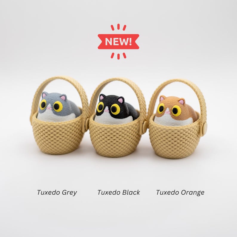 May include: Three small figurines of cats with large yellow eyes, each sitting in a beige woven basket. The cats are colored grey, black, and orange. Above the baskets is a red banner with the text NEW!. Below are the labels Tuxedo Grey, Tuxedo Black, and Tuxedo Orange.
