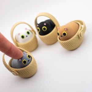 May include: Four small, textured figurines of cats with large yellow eyes and pink ears, each sitting inside a miniature beige woven basket with a handle. The cats are colored white, black, tan, and gray, and they have a rounded, compact shape.
