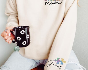 May include: A beige sweatshirt with the text "AUTISM mom" in rainbow colors and a heart. The left sleeve has a rainbow puzzle piece with the name "James" written underneath.