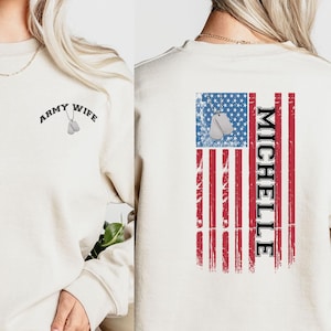 May include: A beige sweatshirt with the text "Army Wife" and a small image of dog tags on the left side. The back of the sweatshirt features a distressed American flag with the text "Michelle" vertically down the right side.