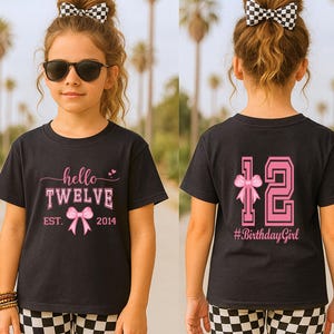 May include: Black t-shirt with the text "hello TWELVE" in pink, a bow graphic, and the year 2014. The back of the shirt has a pink "12" with a bow and the hashtag "#BirthdayGirl". The person is wearing checkered shorts.