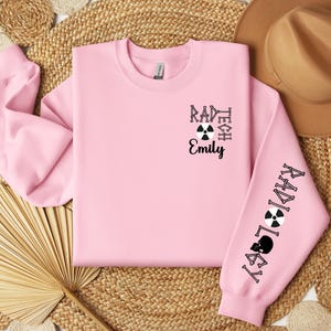 May include: Pink sweatshirt with black text that reads "Rad Tech Emily" and a black and white radiation symbol. The right sleeve has the word "Radiology" written vertically in black with a black and white radiation symbol.