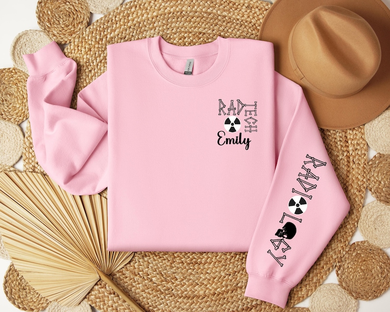 May include: Pink sweatshirt with black text that reads "Rad Tech Emily" and a black and white radiation symbol. The right sleeve has the word "Radiology" written vertically in black with a black and white radiation symbol.