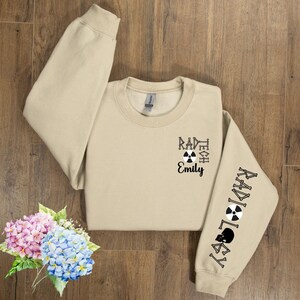 May include: A tan sweatshirt with the text "Rad Tech Emily" and a black and white radiation symbol on the front. The right sleeve has the text "Radiology" with a black and white radiation symbol and a skull and crossbones.