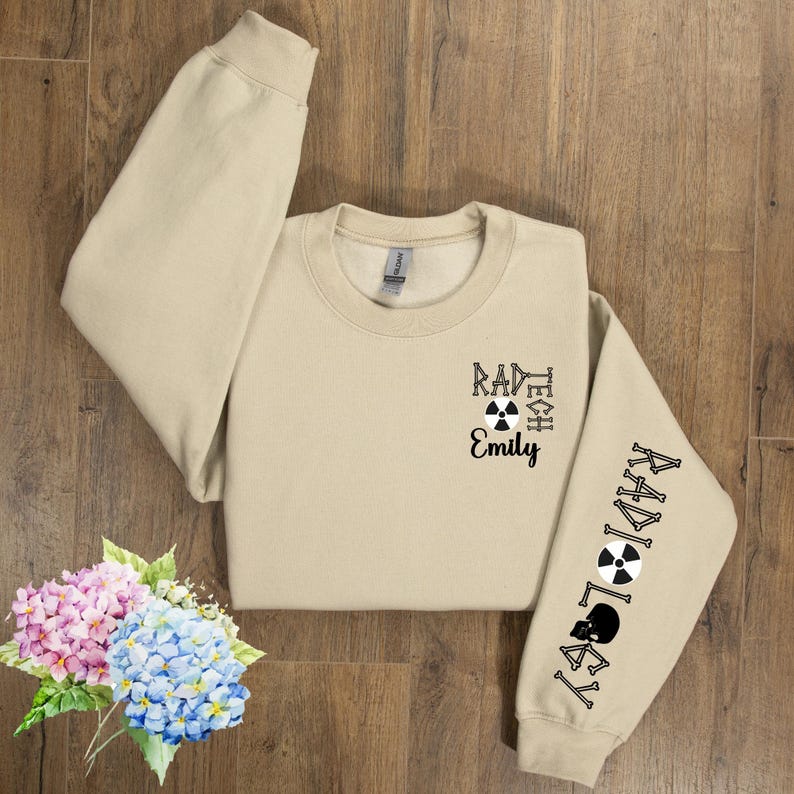 May include: A tan sweatshirt with the text "Rad Tech Emily" and a black and white radiation symbol on the front. The right sleeve has the text "Radiology" with a black and white radiation symbol and a skull and crossbones.