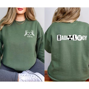 May include: Green sweatshirt with a white graphic of two skeleton hands holding a heart with the word "Radiology" written below. The back of the sweatshirt has the word "RADIOLOGY" written in white with a black radiation symbol and a black and white image of a rib cage.