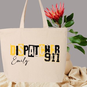 May include: A beige canvas tote bag with black text that reads "Dispatcher 911" and "Emily". The bag has a yellow background behind the text.