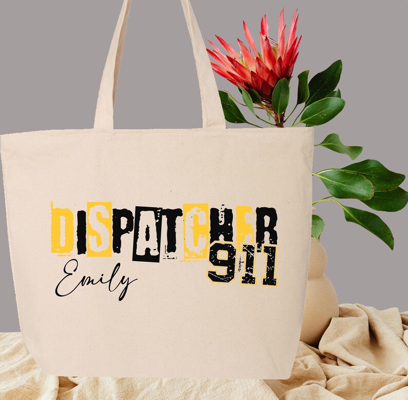 May include: A beige canvas tote bag with black text that reads "Dispatcher 911" and "Emily". The bag has a yellow background behind the text.