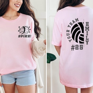 May include: Pink volleyball t-shirt with a graphic of a volleyball player spiking the ball on the front and the text "YOUR TEAM" and a volleyball with the text "EMILY #25" on the back.
