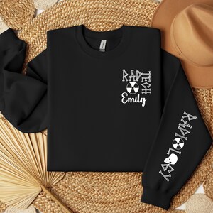 May include: Black sweatshirt with white text that reads "RAD TECH" and "RADIOLOGY" with a skull and crossbones symbol on the sleeves. The text "Emily" is centered on the front of the sweatshirt.