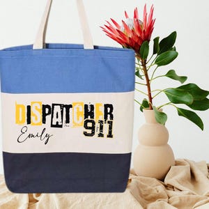 May include: A blue, white, and navy blue canvas tote bag with the text "DISPATCHER 911" in black and yellow letters. The name "Emily" is written in black below the text.