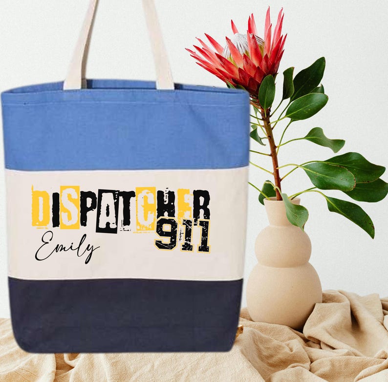 May include: A blue, white, and navy blue canvas tote bag with the text "DISPATCHER 911" in black and yellow letters. The name "Emily" is written in black below the text.
