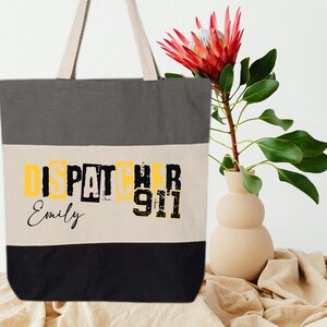 May include: A gray, beige, and black canvas tote bag with the text "DISPATCHER 911" in yellow and black letters. The name "Emily" is written in cursive below the text.