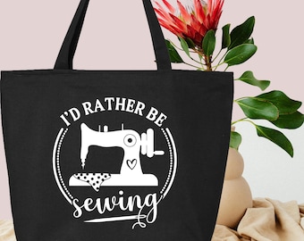 May include: Black tote bag with white text and a sewing machine graphic. The text reads "I'd rather be sewing". The bag has two black handles and is made of a sturdy material. A red flower and green leaves are in the background.
