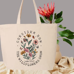 May include: Cream-colored tote bag with a floral design and the text "CONSIDER HOW THE WILDFLOWERS GROW" in a circular arrangement. The design includes pink, yellow, and orange flowers, butterflies, and the Bible verse Luke 12:27.