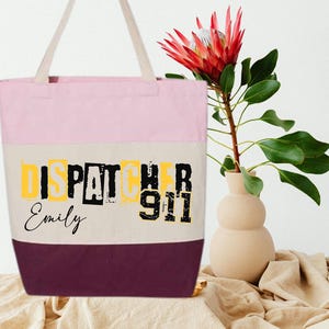 May include: Pink, beige, and burgundy tote bag with the text "Dispatcher 911" in black and yellow letters. The name "Emily" is written in cursive below the text.