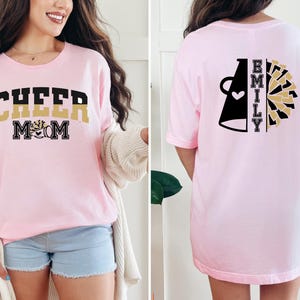Pu&ograve; includere: Maglietta rosa con il testo "CHEER MOM" in nero e oro sul davanti e un megafono nero con un cuore e il testo "EMILY" in oro sul retro.