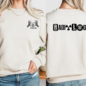 May include: Two cream-colored sweatshirts with black text and graphics. One sweatshirt has a skeleton hand heart graphic with the text "Kimberly". The other sweatshirt has the text "RADIOLOGY" with a radiation symbol, the text "RAD TECH", and a ribcage graphic.